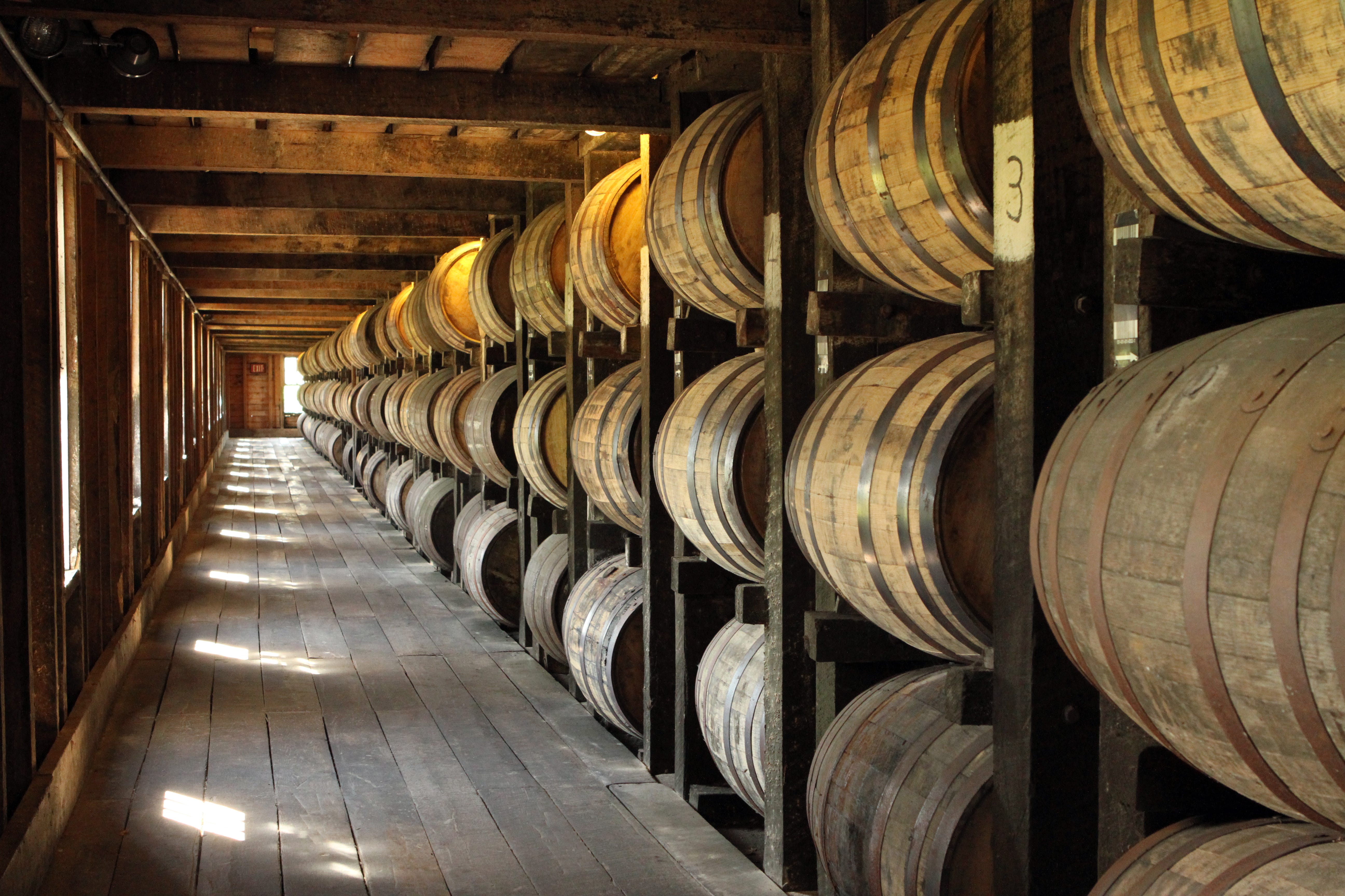 Sun shines through the window of a wooden warehouse. There are rows and rows of bourbon barrels stacked on each other.