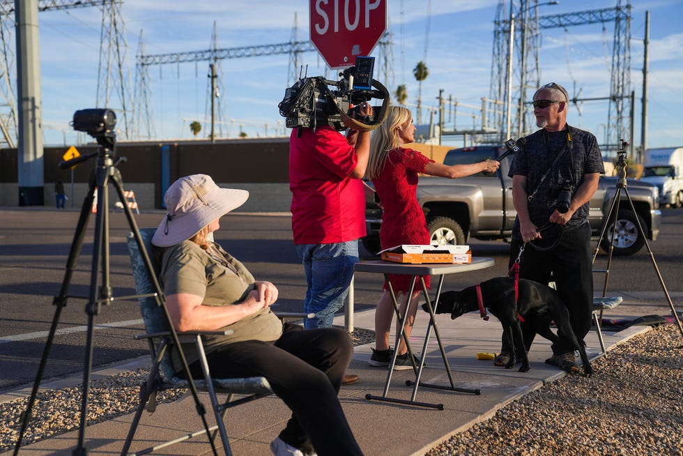 A television news crew interviews people watching, photographing, and recording people as they exit and enter the Maricopa County Tabulation and Elections Center on Oct. 19, 2022, in Phoenix.