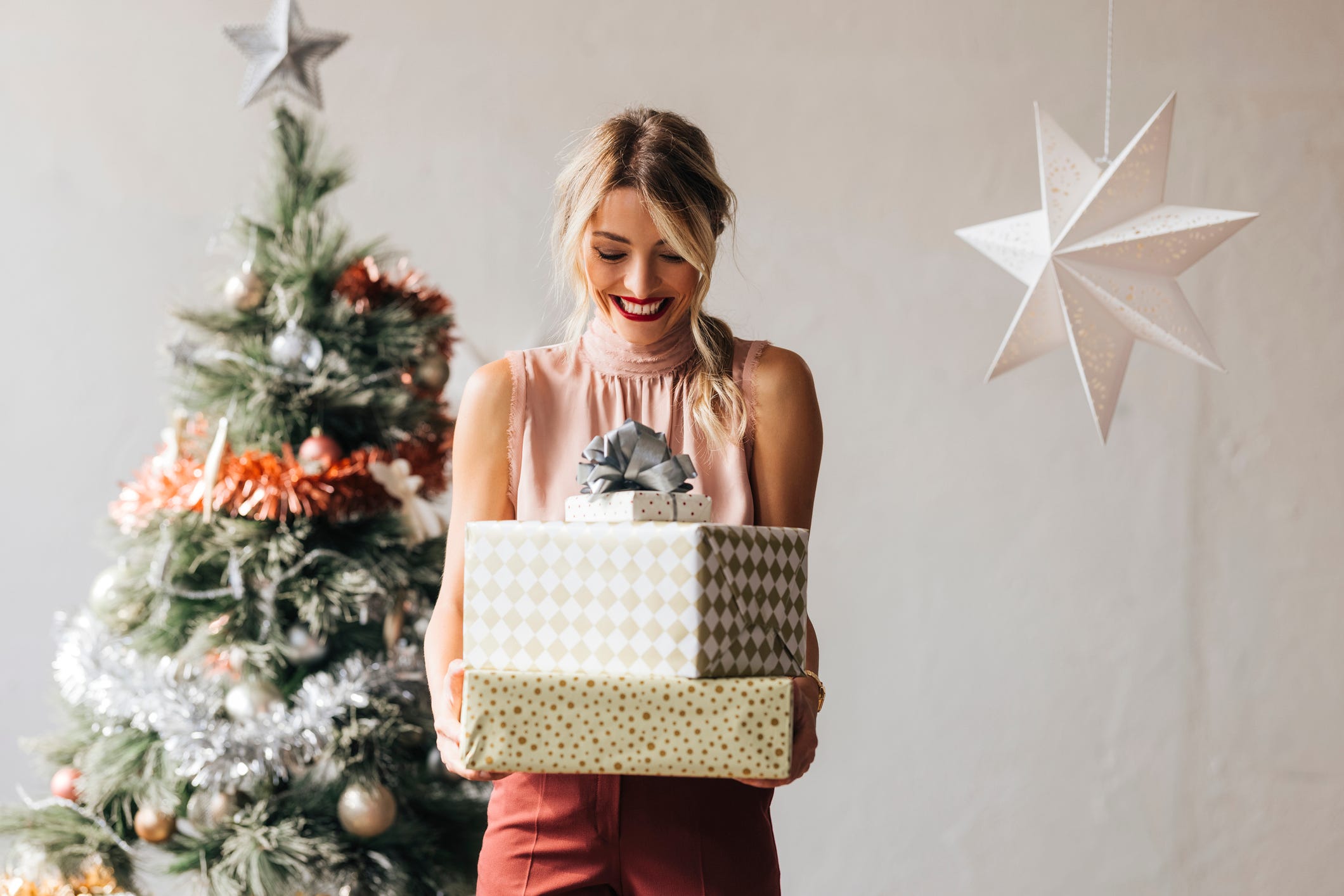 Cheerful smiling female standing next to the decorated Christmas tree and holding wrapped gift boxes in hands