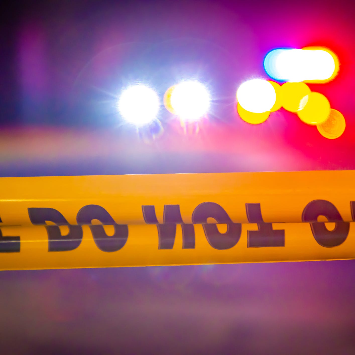 Generic and blurry police lights from a cruiser vehicle and car headlights behind yellow crime scene do not cross caution tape at night.