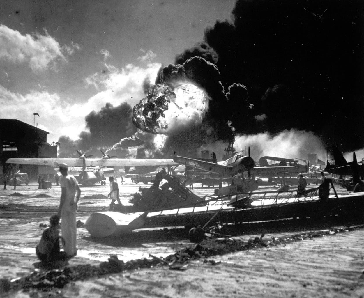 U.S. Navy sailors stand among wrecked airplanes at Ford Island Naval Air Station as they watch the explosion of the USS Shaw in the background, during the Japanese surprise attack on Pearl Harbor.