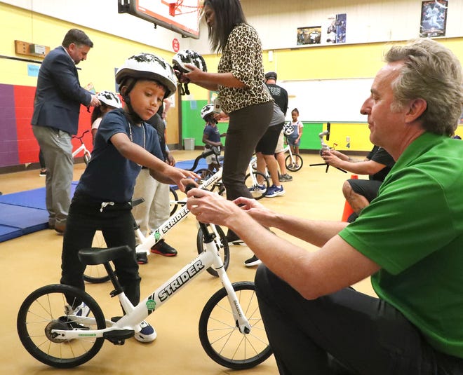 Ryan McFarland, founder and board member of the Strider Education Foundation and All Kids Bike, helps Abraham Torres prepare to ride a Strider bike, Tuesday, March 7, 2023, as Gene Crouch, CEO of AMA Pro Racing, and Karen Troutman, principal of Palm Terrace Elementary, help other students get ready for a ride at the school.