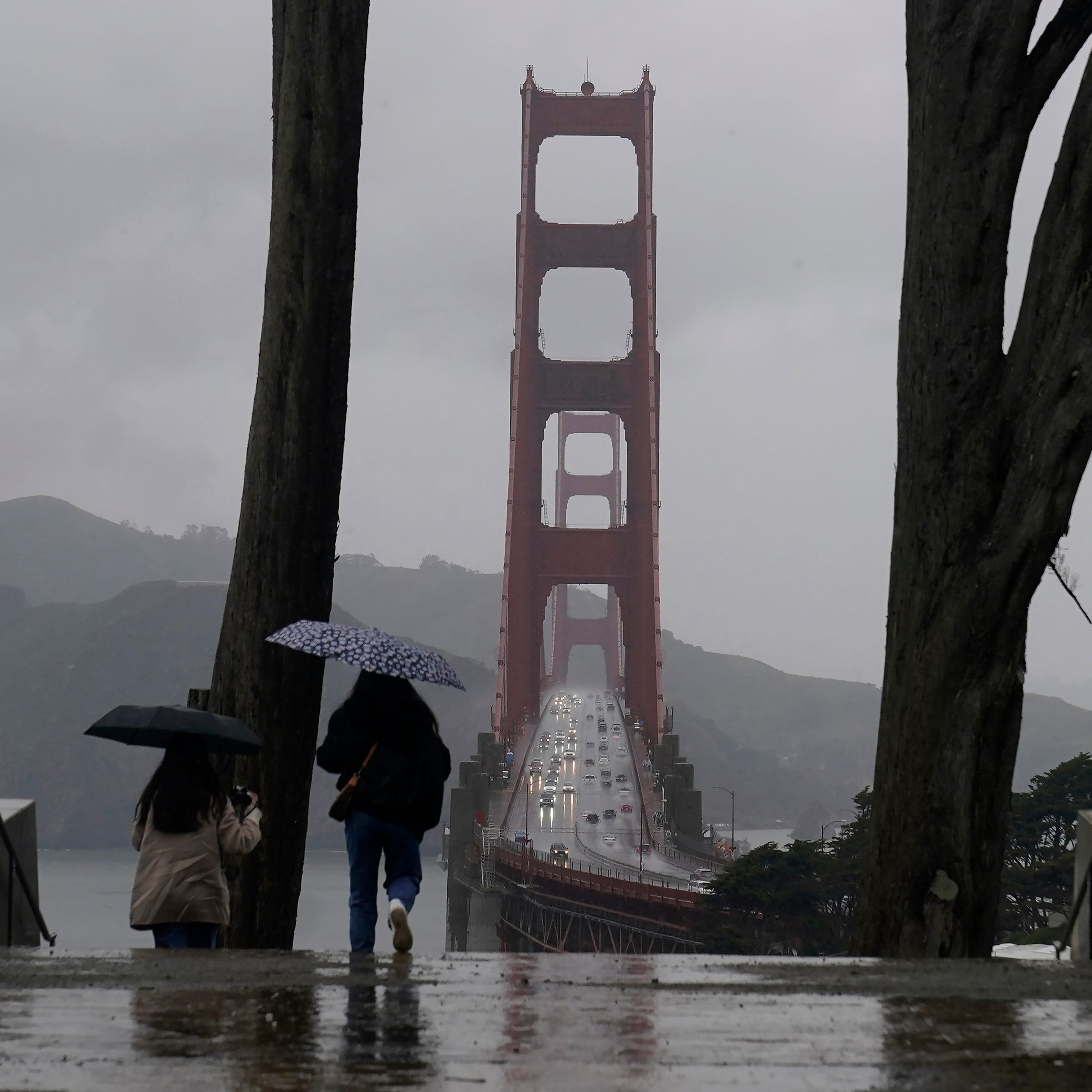 Traffic moves on the Golden Gate Bridge as people carry umbrellas while walking down a path at the Golden Gate Overlook in San Francisco, March 9, 2023. California is bracing for the arrival of an atmospheric river that forecasters warn will bring heavy rain, strong winds, thunderstorms and the threat of flooding even as the state is still digging out from earlier storms.