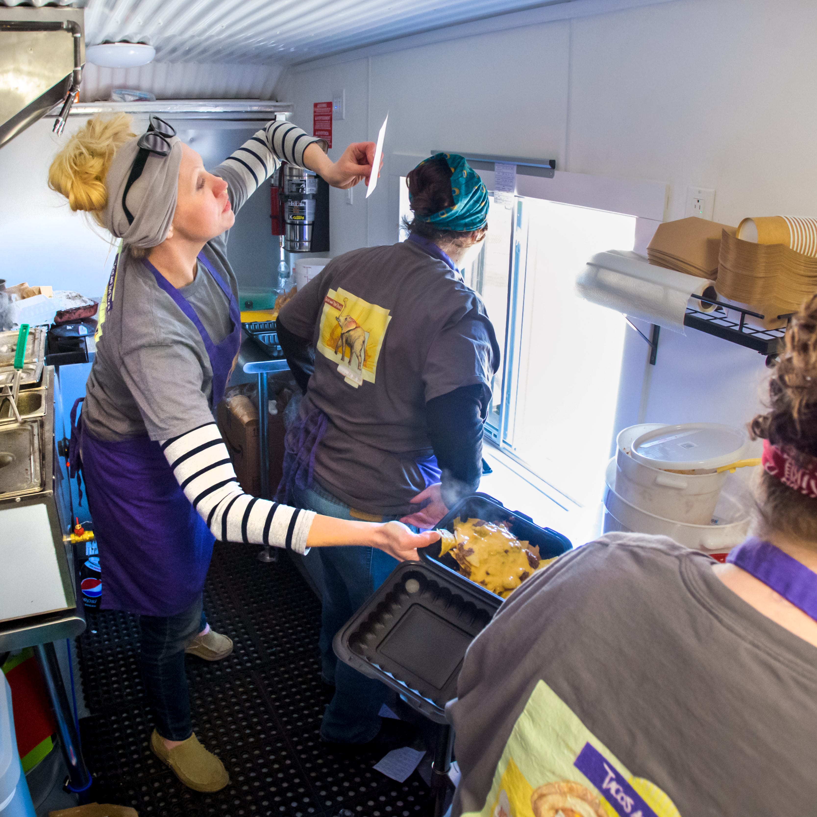 Laura Lytle checks a ticket while handing over an order of nachos to business partner and food prepper Devin Wyman as Kat Ashdown serves customers at the window of the Tacos and Thai food truck during a visit to Illinois CancerCare off Route 91 in North Peoria.